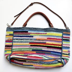May include: A large, colorful crochet tote bag with a brown leather handle and a detachable shoulder strap. The bag features a patchwork design with horizontal stripes in various colors, including blue, yellow, pink, and black. The bag is approximately 15 inches wide.