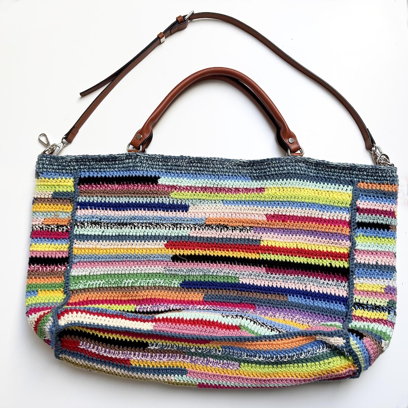 May include: A large, colorful crochet tote bag with a brown leather handle and a detachable shoulder strap. The bag features a patchwork design with horizontal stripes in various colors, including blue, yellow, pink, and black. The bag is approximately 15 inches wide.