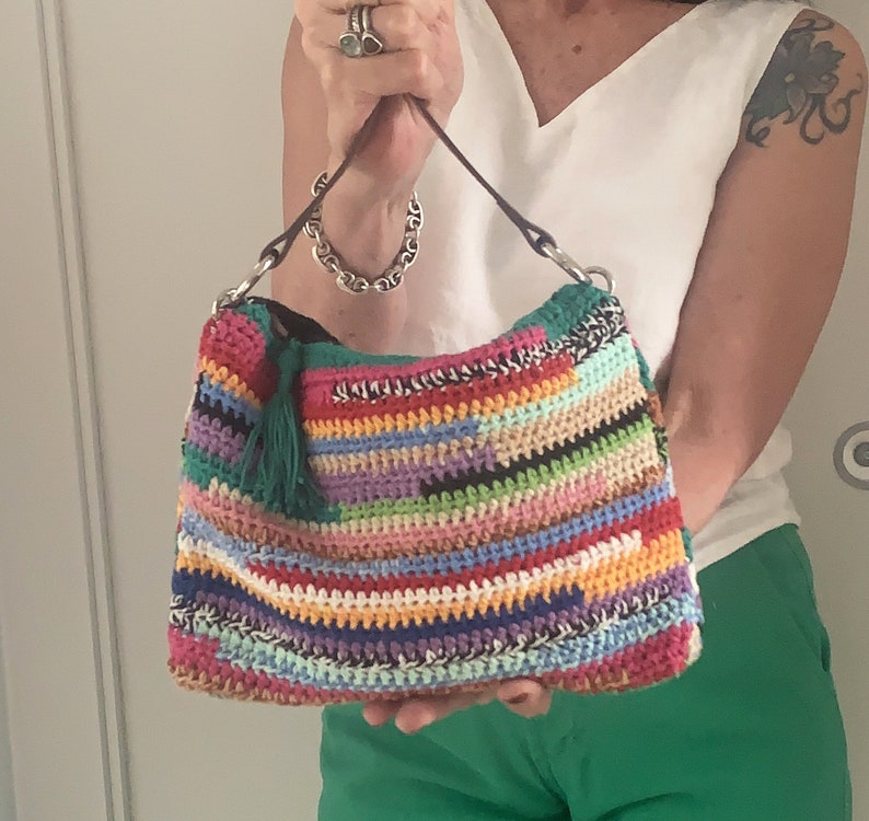 May include: A colorful crocheted handbag with a green tassel. The bag has a brown leather strap and is made with a variety of different colored yarns.