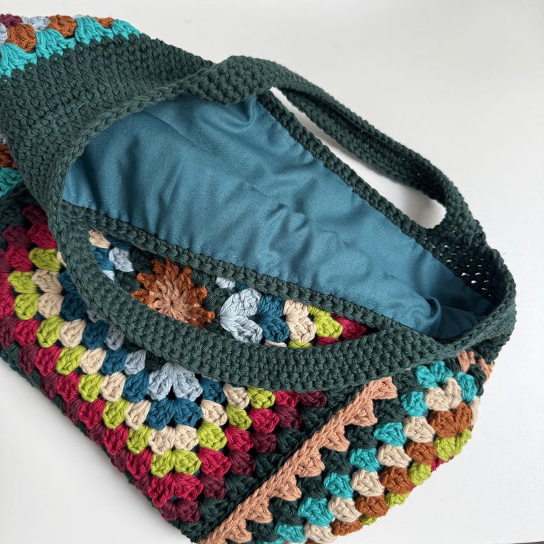 Puede incluir: Un bolso tote de crochet hecho a mano con un dise&ntilde;o de cuadrados de la abuela colorido. El bolso presenta un exterior verde oscuro con un forro interior azul verdoso. Las asas y el ribete tambi&eacute;n son de color verde oscuro.