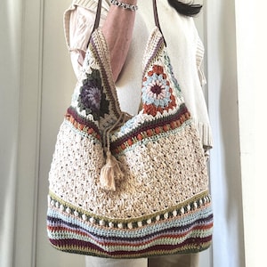 May include: A cream-colored crochet tote bag with brown leather straps. The bag features colorful floral and geometric patterns in shades of purple, orange, green, and blue. A beige tassel hangs from the drawstring closure. The bag is approximately 15 inches wide and 14 inches tall.