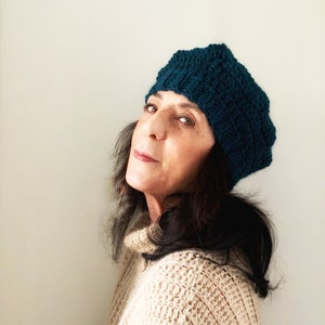 May include: A woman with long dark hair is wearing a teal blue knitted beanie. She is looking off to the side and wearing a beige sweater.
