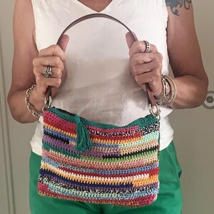May include: A colorful crocheted handbag with a striped pattern and a green tassel. The bag has a brown leather strap and silver hardware.