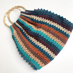 Puede incluir: Un bolso de mano de crochet a rayas con un asa de bambú. El bolso está hecho con una variedad de colores, incluyendo azul verdoso, marrón, naranja y beige. El bolso tiene un borde de crochet.