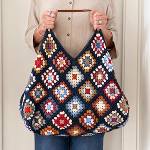 May include: A large, navy blue crochet tote bag with a colorful granny square pattern. The bag features a brown leather handle and is held up by a person. The granny squares are in shades of red, orange, blue, and cream.