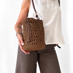 May include: A brown bucket bag crafted from woven straw-like material featuring an open-work crochet pattern. The bag includes a long, thin dark brown leather shoulder strap with a knot detail and a drawstring closure at the top opening.