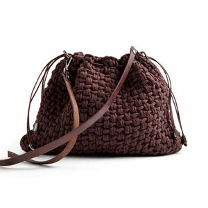 May include: A dark brown woven shoulder bag with a drawstring closure. It has a long, brown leather strap and silver-toned hardware. The bag is a medium size, suitable for everyday use. The bag's dimensions are approximately 30cm x 30cm.