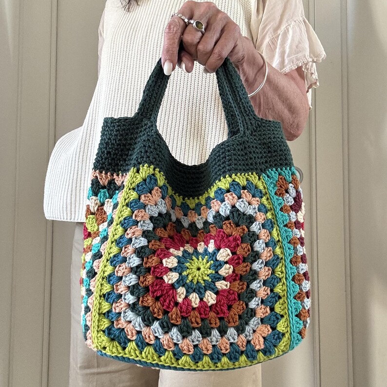 Puede incluir: Un bolso tote de ganchillo hecho a mano y colorido. El bolso presenta un dise&ntilde;o de cuadrados de la abuela con una variedad de colores, incluyendo verde, azul, rojo y naranja. Las asas y el borde superior son de color verde oscuro liso. El bolso es sostenido por una persona.
