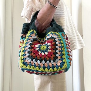 Puede incluir: Un bolso tote de crochet hecho a mano y colorido. El bolso presenta un dise&ntilde;o de cuadrados de la abuela con una variedad de colores, incluyendo rojo, verde, azul y amarillo. El bolso tiene un asa y ribete verde oscuro. El bolso es sostenido por una persona.