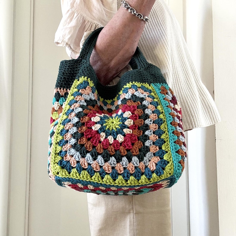 Puede incluir: Un bolso tote de crochet hecho a mano y colorido. El bolso presenta un dise&ntilde;o de cuadrados de la abuela con una variedad de colores, incluyendo rojo, verde, azul y amarillo. El bolso tiene un asa y ribete verde oscuro. El bolso es sostenido por una persona.