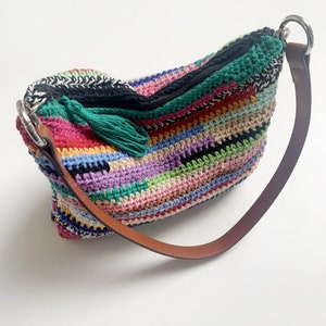 May include: A colorful crocheted shoulder bag with a green tassel and a brown leather strap. The bag has a zipper closure and a silver metal ring.