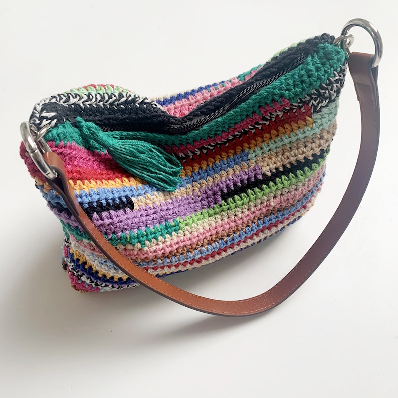 May include: A colorful crocheted shoulder bag with a green tassel and a brown leather strap. The bag has a zipper closure and a silver metal ring.