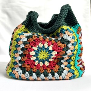 Puede incluir: Bolso tote de crochet hecho a mano con un dise&ntilde;o de cuadrados de la abuela colorido. El bolso presenta un asa y ribete verde oscuro, con una vibrante mezcla de cuadrados rojos, amarillos, azules y marrones. Un accesorio elegante y funcional.