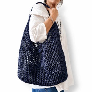 Puede incluir: Un bolso de hombro de ganchillo en color azul marino oscuro con un patrón de red de tejido abierto y un cierre de botón circular único en la parte delantera. El bolso tiene una correa ancha integrada y una silueta holgada y flexible.