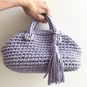 May include: A lavender crocheted handbag with a rounded shape and two handles. The bag features a matching tassel and a decorative bead. The texture of the bag is visible, showcasing the handmade quality.