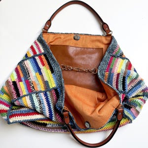May include: A large, open tote bag with a colorful, patchwork crochet exterior. The bag features brown leather handles and trim, with a brown leather interior pocket and a chain closure. The interior lining is a bright orange fabric.