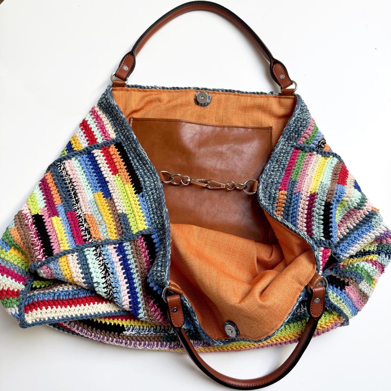May include: A large, open tote bag with a colorful, patchwork crochet exterior. The bag features brown leather handles and trim, with a brown leather interior pocket and a chain closure. The interior lining is a bright orange fabric.