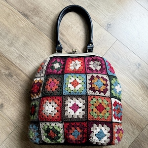 May include: A colorful crocheted handbag with a black leather handle and a brass clasp. The bag is made of squares of different colors, including red, green, blue, and brown.