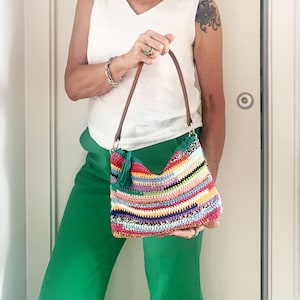 May include: A colorful crocheted shoulder bag with a brown leather strap. The bag has a fringe detail and is made with a variety of bright colors.