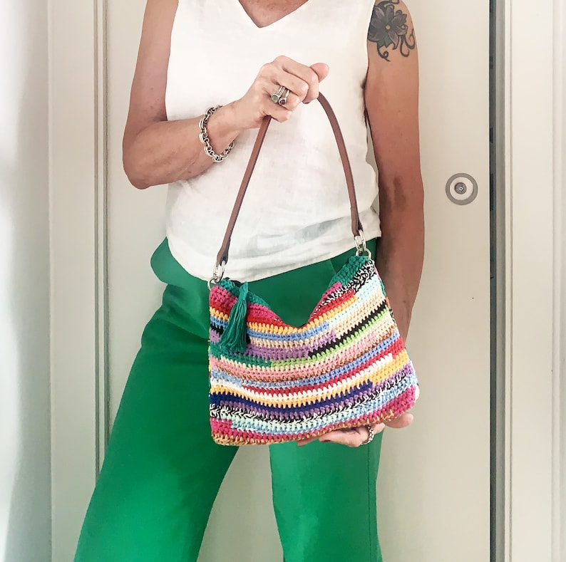 May include: A colorful crocheted shoulder bag with a brown leather strap. The bag has a fringe detail and is made with a variety of bright colors.
