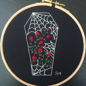 May include: Embroidered hoop art featuring a white coffin with a spiderweb design and red roses. The background is black fabric and the hoop is made of wood. The artist's initials "SM" are embroidered in the bottom right corner.