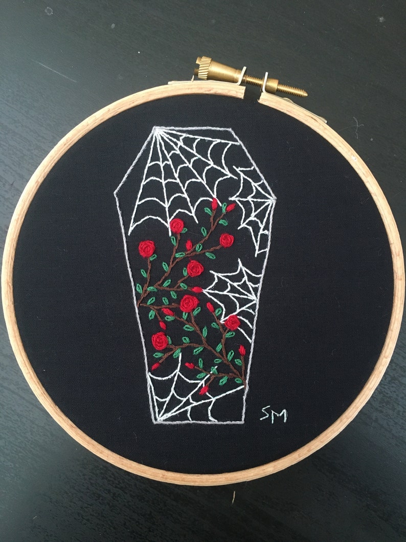 May include: Embroidered hoop art featuring a white coffin with a spiderweb design and red roses. The background is black fabric and the hoop is made of wood. The artist's initials "SM" are embroidered in the bottom right corner.