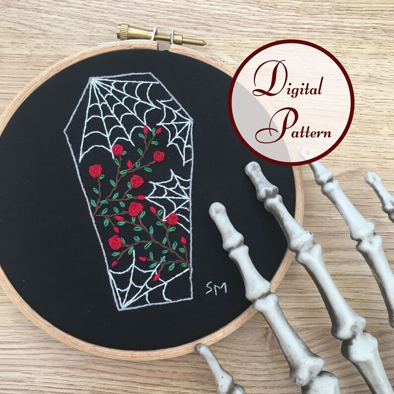 May include: Embroidered design of a white coffin with a spiderweb and red roses on a black fabric background. The design is in a circular embroidery hoop.