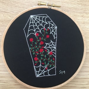 May include: Embroidered hoop art featuring a white coffin with a spiderweb inside and red roses growing around it. The background is black fabric and the hoop is wooden.