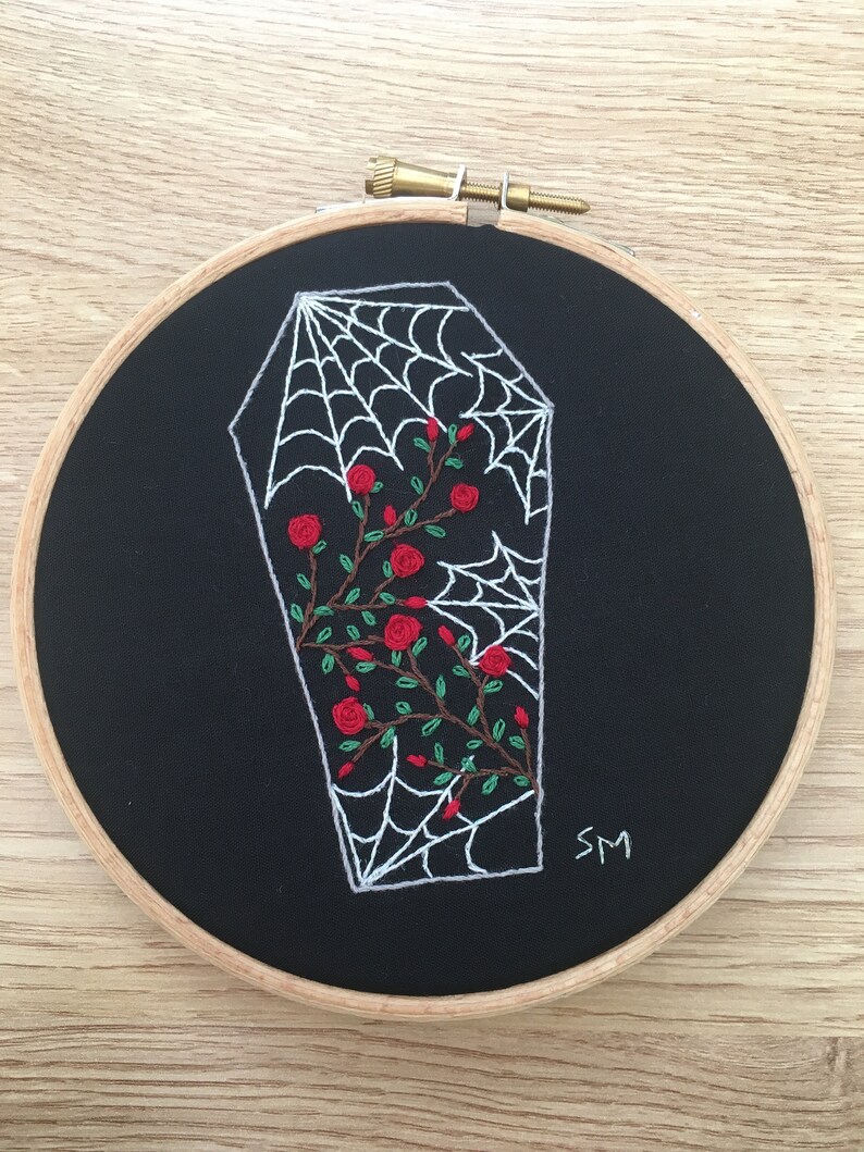 May include: Embroidered hoop art featuring a white coffin with a spiderweb inside and red roses growing around it. The background is black fabric and the hoop is wooden.