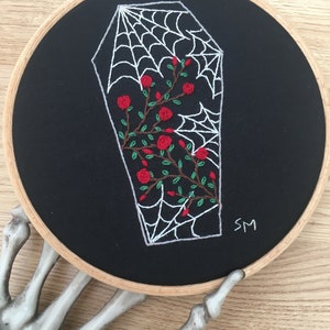 May include: Embroidered hoop art featuring a white coffin with a spiderweb design and red roses inside. The background is black fabric. The hoop is held by a skeleton hand. The artist's initials "SM" are embroidered in the bottom right corner.