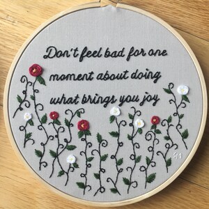 Puede incluir: Arte de bordado en aro con fondo gris y texto en cursiva negra que dice "No te sientas mal por un momento por hacer lo que te trae alegr&iacute;a". El texto est&aacute; rodeado de enredaderas negras con flores rojas y blancas.