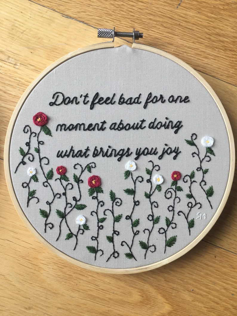 Puede incluir: Arte de bordado en aro con fondo gris y texto en cursiva negra que dice "No te sientas mal por un momento por hacer lo que te trae alegr&iacute;a". El texto est&aacute; rodeado de enredaderas negras con flores rojas y blancas.