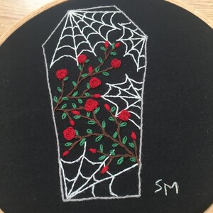 May include: Embroidered hoop art featuring a white spiderweb over a black coffin with red roses and green leaves. The hoop is made of wood and has a black fabric background. The artist's initials "SM" are embroidered in the bottom right corner.