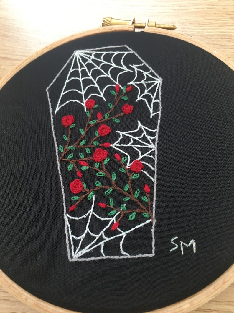 May include: Embroidered hoop art featuring a white spiderweb over a black coffin with red roses and green leaves. The hoop is made of wood and has a black fabric background. The artist's initials "SM" are embroidered in the bottom right corner.