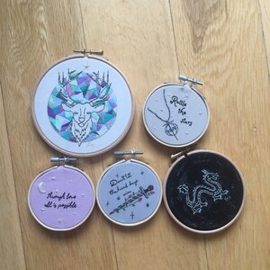 Puede incluir: Cinco aros bordados con diferentes dise&ntilde;os y lemas. Los aros son blancos, morado claro, gris, negro y blanco. Los lemas son "No dejes que los d&iacute;as dif&iacute;ciles te ganen", "Agita las estrellas", "A trav&eacute;s del amor todo es posible", y un dise&ntilde;o de un drag&oacute;n. El &uacute;ltimo aro tiene un dise&ntilde;o geom&eacute;trico de una cabeza de ciervo.