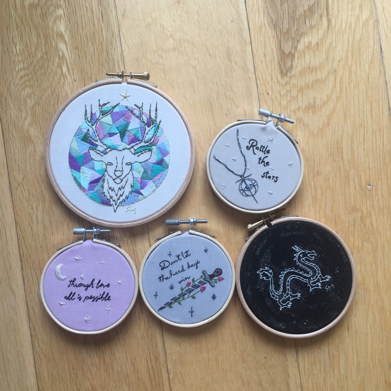 Puede incluir: Cinco aros bordados con diferentes dise&ntilde;os y lemas. Los aros son blancos, morado claro, gris, negro y blanco. Los lemas son "No dejes que los d&iacute;as dif&iacute;ciles te ganen", "Agita las estrellas", "A trav&eacute;s del amor todo es posible", y un dise&ntilde;o de un drag&oacute;n. El &uacute;ltimo aro tiene un dise&ntilde;o geom&eacute;trico de una cabeza de ciervo.