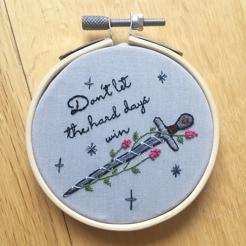 Puede incluir: Arte de bordado en forma de aro con fondo gris y una espada negra con una joya roja. La espada est&aacute; rodeada de enredaderas verdes y flores rosas. El texto "Don't let the hard days win" est&aacute; bordado en hilo negro.
