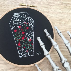 May include: Embroidered hoop art featuring a white coffin with a spiderweb design and red roses. The background is black and the hoop is wooden.