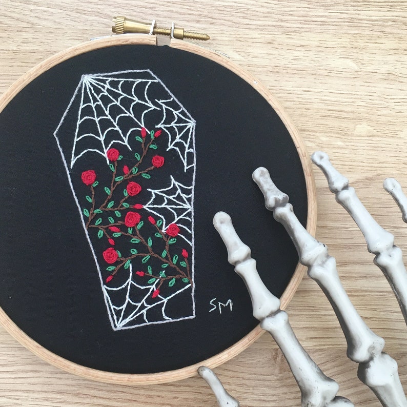 May include: Embroidered hoop art featuring a white coffin with a spiderweb design and red roses. The background is black and the hoop is wooden.