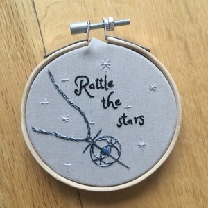 Puede incluir: Arte de bordado en aro con fondo gris y el texto "Rattle the stars" en cursiva negra. Un dise&ntilde;o en forma de estrella azul y negro est&aacute; cosido en el centro del aro.