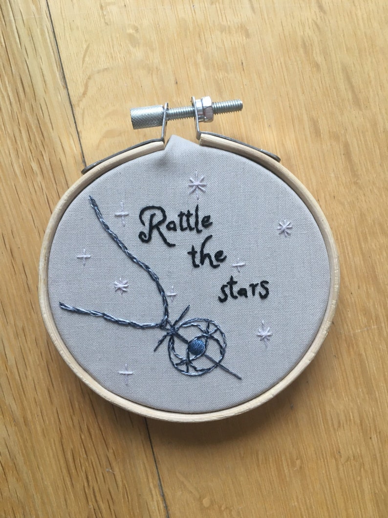 Puede incluir: Arte de bordado en aro con fondo gris y el texto "Rattle the stars" en cursiva negra. Un dise&ntilde;o en forma de estrella azul y negro est&aacute; cosido en el centro del aro.