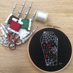 May include: A black embroidery hoop with a white spiderweb design inside a coffin shape. The spiderweb has red roses growing inside it. The hoop is on a wooden surface with a skeleton hand holding embroidery floss.