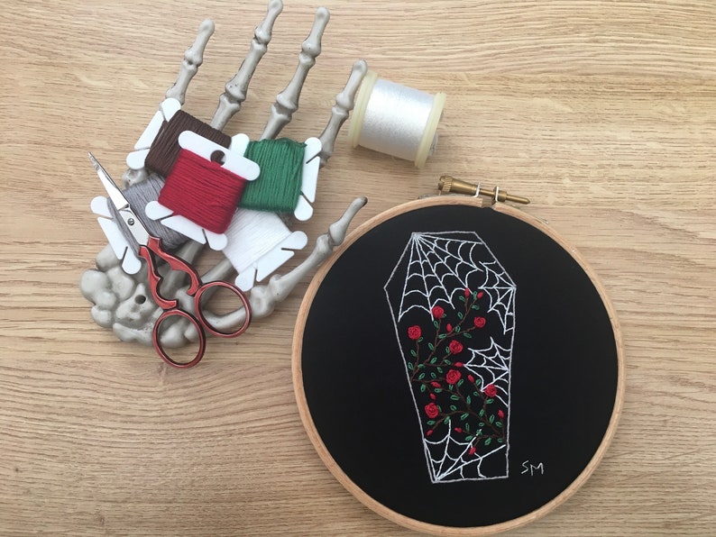 May include: A black embroidery hoop with a white spiderweb design inside a coffin shape. The spiderweb has red roses growing inside it. The hoop is on a wooden surface with a skeleton hand holding embroidery floss.