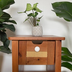 May include: A wooden nightstand with a drawer and a lower shelf. The drawer has a white knob and a woven pattern on the front. A small potted plant sits on top of the nightstand.