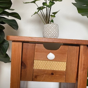 Könnte beinhalten: Ein Nachttisch aus Holz mit einer Schublade und einem Regal. Die Schublade hat einen Muschel-Griff und ein gewebtes Muster auf der Vorderseite. Eine Topfpflanze steht auf dem Nachttisch.