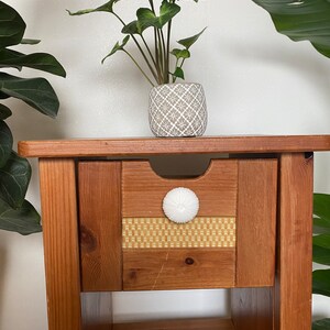 May include: A wooden nightstand with a single drawer and a lower shelf. The drawer has a white knob and a woven pattern on the front. A small potted plant sits on top of the nightstand.