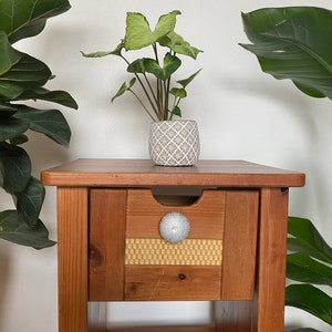 May include: A wooden end table with a single drawer and a lower shelf. The drawer has a white knob with a textured design. A potted plant with green leaves sits on the top of the table.