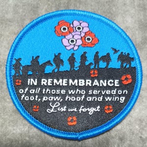 May include: A circular embroidered patch with a blue border. The patch features a black silhouette of soldiers, horses, and birds against a blue background. Text reads "IN REMEMBRANCE" and "List we forget". Red and purple flowers are at the top.