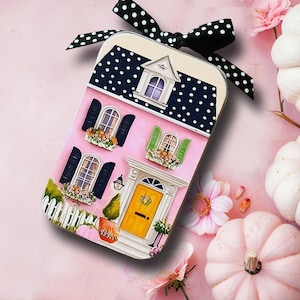 May include: A rectangular metal tin featuring a printed illustration of a pink house with a yellow door, black and white polka dot roof, and a matching black and white polka dot ribbon tied to the top, surrounded by white pumpkins and pink flowers.