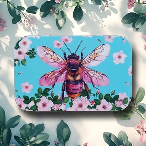 May include: A rectangular, light blue tin with a watercolor illustration of a bee with pink and white wings. The bee is surrounded by pink flowers and green leaves. The tin is a decorative item.